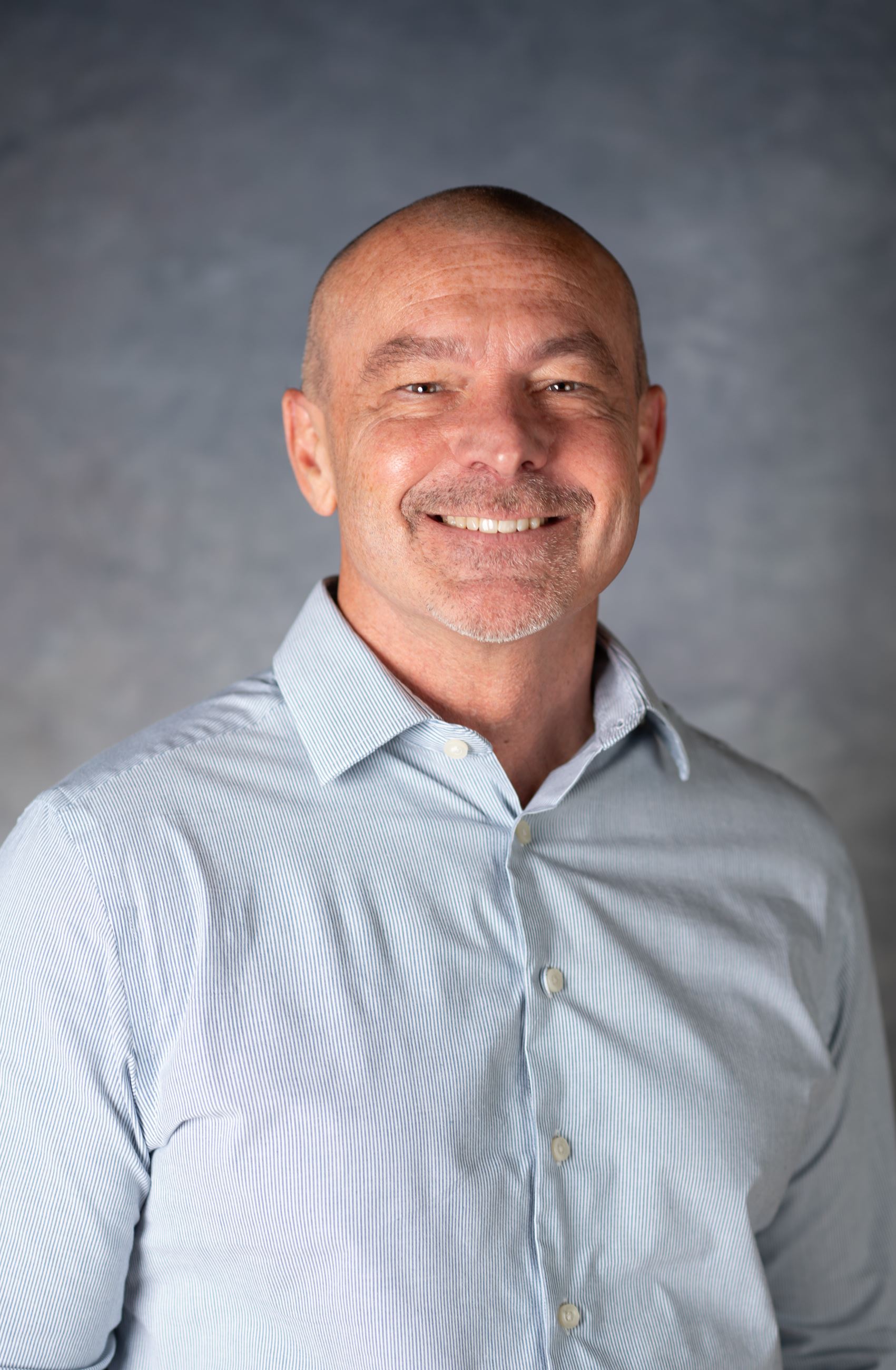 Bald man with blue collared shirt smiles wide directly at camera.